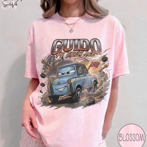 May include: A light pink t-shirt featuring a cartoon image of Guido from the movie Cars. The graphic includes the text "Guido Pit Crew Ace" in a metallic font, with a blue car and a background of tools and smoke.