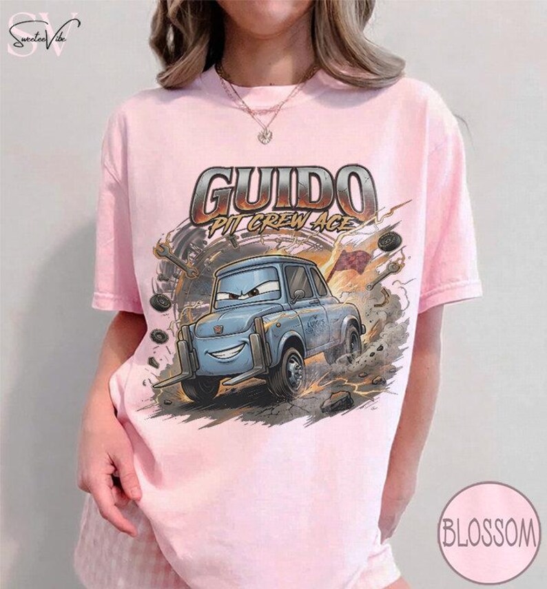 May include: A light pink t-shirt featuring a cartoon image of Guido from the movie Cars. The graphic includes the text "Guido Pit Crew Ace" in a metallic font, with a blue car and a background of tools and smoke.