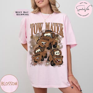 K&ouml;nnte beinhalten: Hellrosa T-Shirt mit einem braunen Aufdruck der Figur Abschleppwagen Mater aus dem Film Cars. Der Text "TOW MATER" befindet sich &uuml;ber der Grafik. Das T-Shirt hat kurze &Auml;rmel und eine lockere Passform.