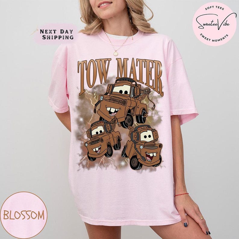 K&ouml;nnte beinhalten: Hellrosa T-Shirt mit einem braunen Aufdruck der Figur Abschleppwagen Mater aus dem Film Cars. Der Text "TOW MATER" befindet sich &uuml;ber der Grafik. Das T-Shirt hat kurze &Auml;rmel und eine lockere Passform.
