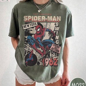 May include: Olive green t-shirt featuring a vintage Spider-Man graphic. The design includes the text "The Amazing Spider-Man," "New York," "Swing," "Spring into Action," and the year "1962." The shirt has a distressed, retro aesthetic.