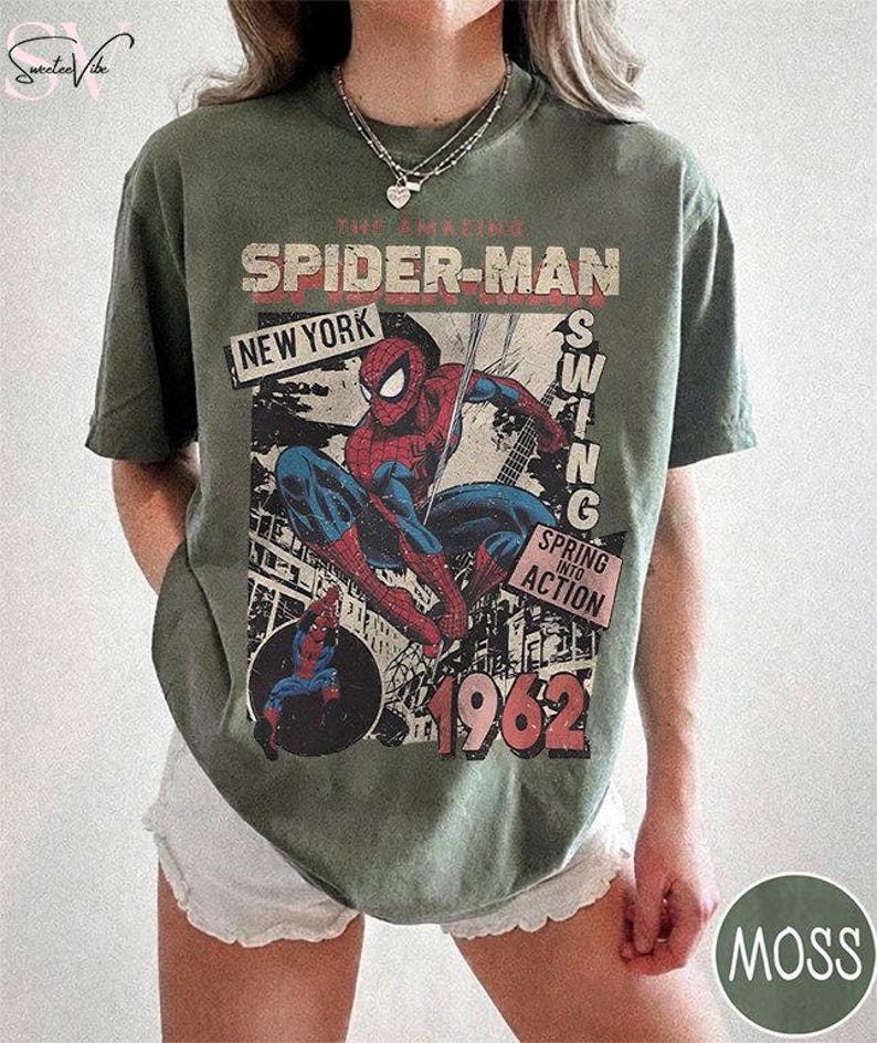 May include: Olive green t-shirt featuring a vintage Spider-Man graphic. The design includes the text "The Amazing Spider-Man," "New York," "Swing," "Spring into Action," and the year "1962." The shirt has a distressed, retro aesthetic.