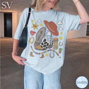 May include: A light blue t-shirt featuring a graphic print of a brown cowboy hat, a cow-print boot with the name Taylor, a sheriff badge, a red bandana, a lasso, a horseshoe, a cactus, and building blocks with the letters T, S, and the number 13.