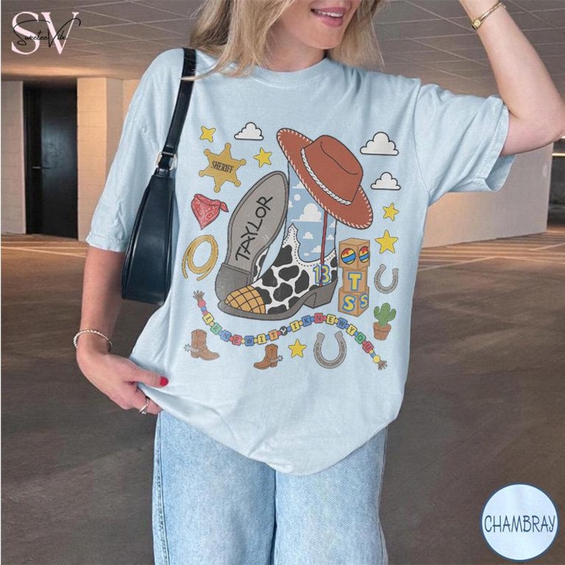May include: A light blue t-shirt featuring a graphic print of a brown cowboy hat, a cow-print boot with the name Taylor, a sheriff badge, a red bandana, a lasso, a horseshoe, a cactus, and building blocks with the letters T, S, and the number 13.