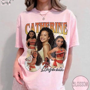 K&ouml;nnte beinhalten: Ein hellrosa T-Shirt mit kurzen &Auml;rmeln und einem Grafikdruck, der den Namen CATHERINE in gro&szlig;en orangefarbenen Buchstaben zeigt. Das Design enth&auml;lt Illustrationen der Figur Moana, ein kleines Schwein und ein zentrales Foto einer Person mit lockigem Haar.