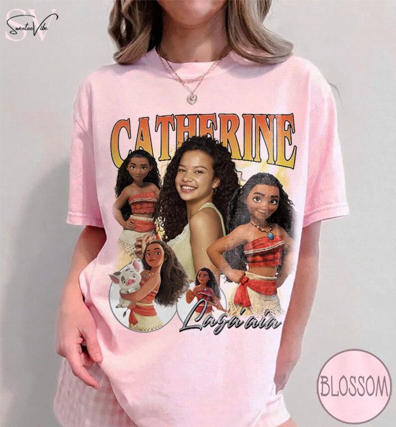 K&ouml;nnte beinhalten: Ein hellrosa T-Shirt mit kurzen &Auml;rmeln und einem Grafikdruck, der den Namen CATHERINE in gro&szlig;en orangefarbenen Buchstaben zeigt. Das Design enth&auml;lt Illustrationen der Figur Moana, ein kleines Schwein und ein zentrales Foto einer Person mit lockigem Haar.