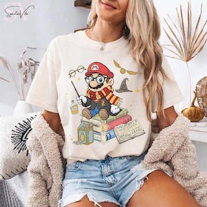May include: Cream-colored t-shirt featuring a cartoon Mario character dressed as a wizard, wearing glasses and a scarf, sitting on books. The design includes a wand, potion bottle, and a golden snitch. The text "Sweetee Vibe" is in the upper left corner.