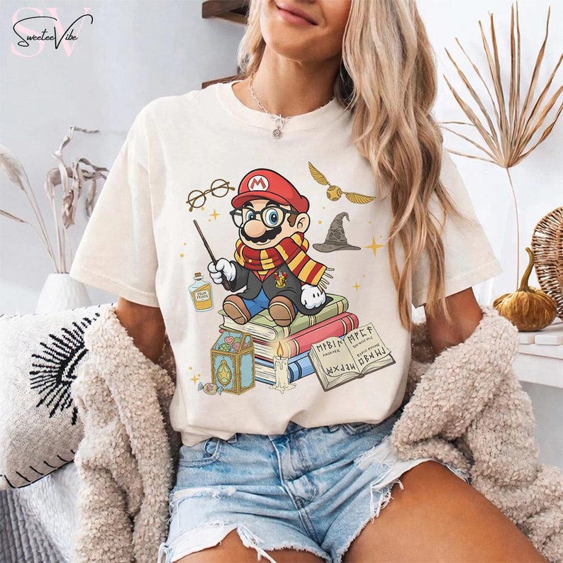 May include: Cream-colored t-shirt featuring a cartoon Mario character dressed as a wizard, wearing glasses and a scarf, sitting on books. The design includes a wand, potion bottle, and a golden snitch. The text "Sweetee Vibe" is in the upper left corner.