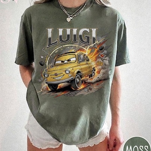 May include: Olive green t-shirt featuring the name "LUIGI" above a cartoon image of a yellow car with a smiling face. The car is depicted in motion with flames and tire tracks. The shirt has a crew neck and short sleeves.