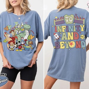 Op de afbeelding: Blauw spijkerbroekkleurig T-shirt met een afbeelding van Toy Story-personages op de voorkant. De achterkant toont de zin "To Infinity and Beyond" in kleurrijke letters, met raket- en steraccenten. Het shirt is een comfort colors-stijl.