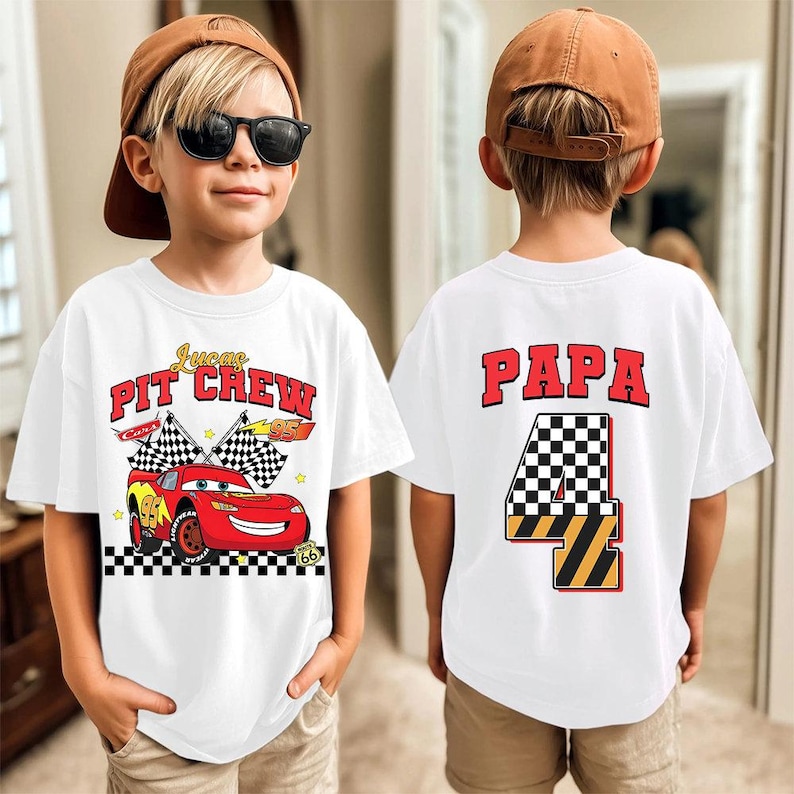 May include: White t-shirt with a red race car graphic and the text "Lucas PIT CREW" on the front. The back of the shirt has the word "PAPA" and the number "4" in a checkered flag design. The shirt is worn by a child.