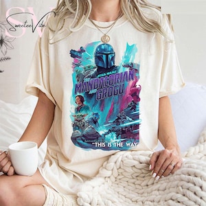 May include: Cream-colored t-shirt featuring a vibrant graphic from "The Mandalorian and Grogu." The design showcases the Mandalorian, Grogu, and other characters in a colorful, action-packed scene. Text on the shirt reads "THIS IS THE WAY."