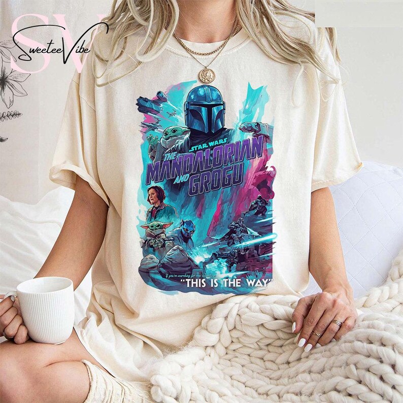 May include: Cream-colored t-shirt featuring a vibrant graphic from "The Mandalorian and Grogu." The design showcases the Mandalorian, Grogu, and other characters in a colorful, action-packed scene. Text on the shirt reads "THIS IS THE WAY."