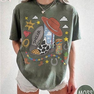 May include: A moss green t-shirt featuring a graphic print of a cowboy boot with a cow print pattern, a brown cowboy hat, a sheriff badge, alphabet blocks, and a beaded string. The boot sole is labeled with the name Taylor and the number 13.