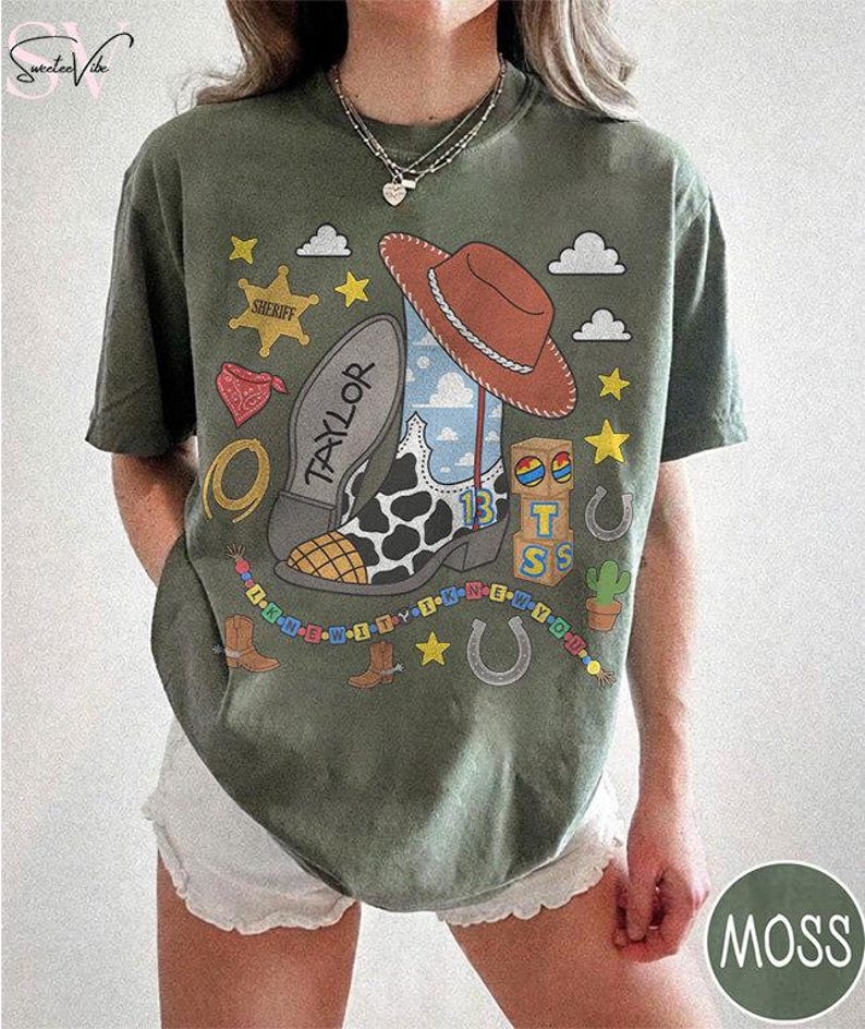 May include: A moss green t-shirt featuring a graphic print of a cowboy boot with a cow print pattern, a brown cowboy hat, a sheriff badge, alphabet blocks, and a beaded string. The boot sole is labeled with the name Taylor and the number 13.
