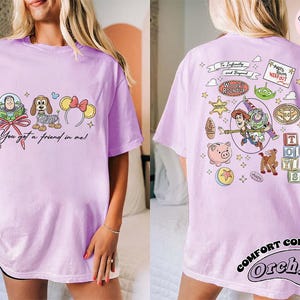May include: A light purple t-shirt featuring Toy Story characters. The front displays Buzz Lightyear, a dog, and Minnie Mouse ears with the text "You got a friend in me!". The back has more characters and the text "To Infinity and Beyond".