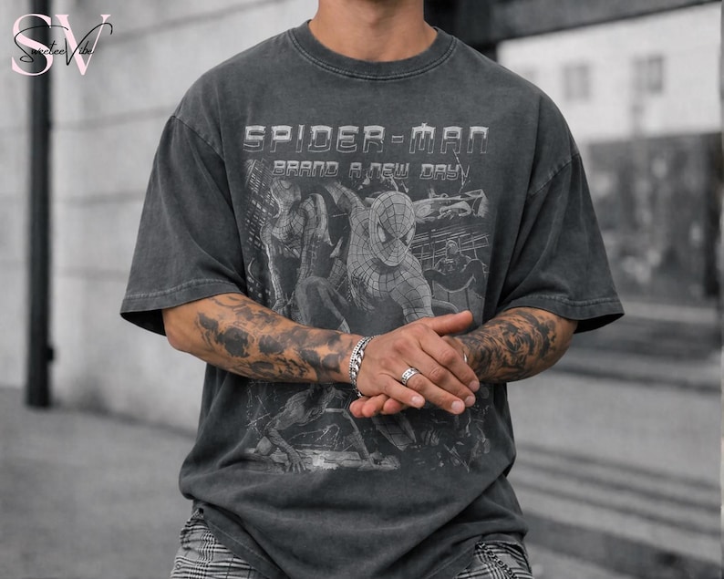 May include: A charcoal gray short-sleeve t-shirt featuring a graphic print of the comic book character Spider-Man. The text "SPIDER-MAN" and "BRAND A NEW DAY" is printed in white above the character illustration on the front of the garment.