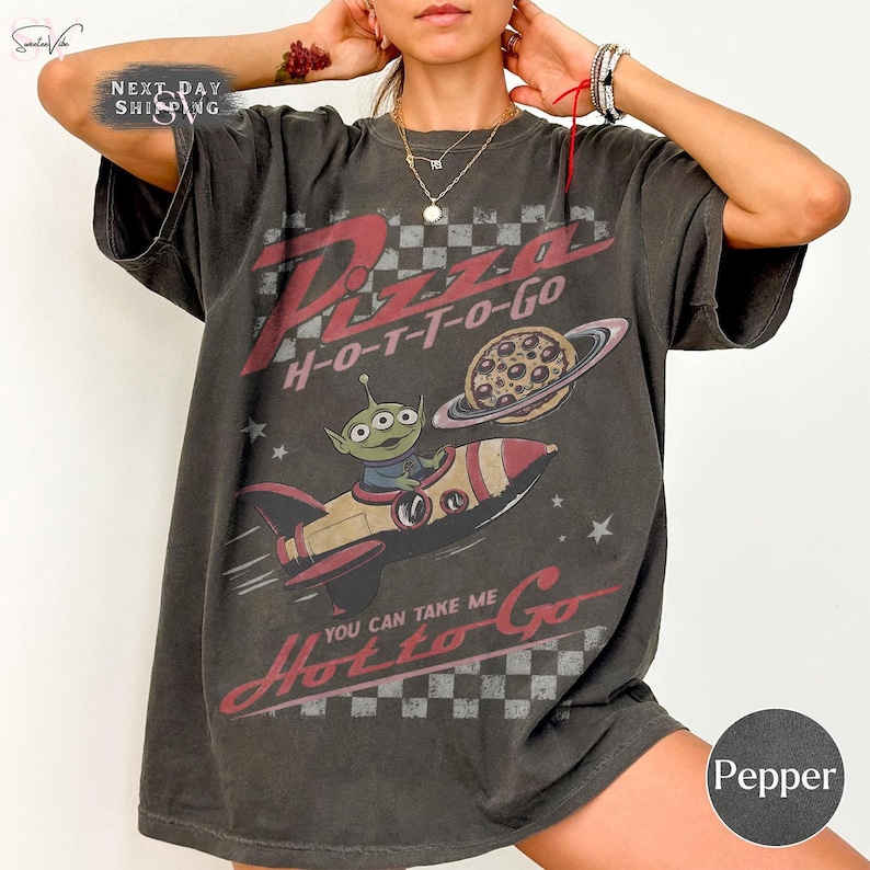 May include: Pepper-colored t-shirt with a distressed graphic. The design features the words "Pizza H-O-T-T-O-Go" in red, a cartoon alien in a rocket, and a pizza with a planet ring. The text "You can take me Hotto Go" is also included.