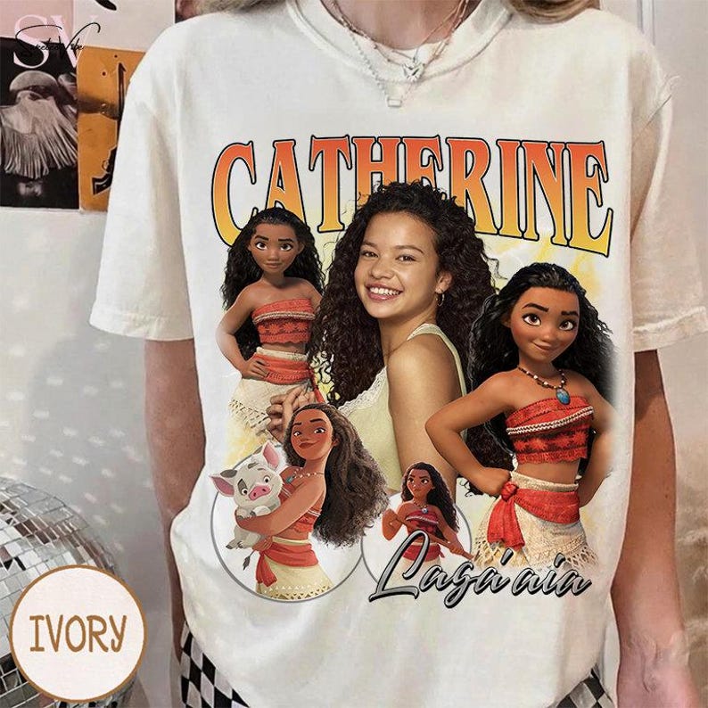 K&ouml;nnte beinhalten: Ein cremefarbenes Kurzarm-T-Shirt mit einem gro&szlig;en Grafikdruck, der den Namen CATHERINE in fetten orangefarbenen Buchstaben zeigt. Das Design enth&auml;lt Illustrationen der Figur Moana, eines kleinen Schweins und ein zentrales Portr&auml;t einer Person mit lockigem Haar.