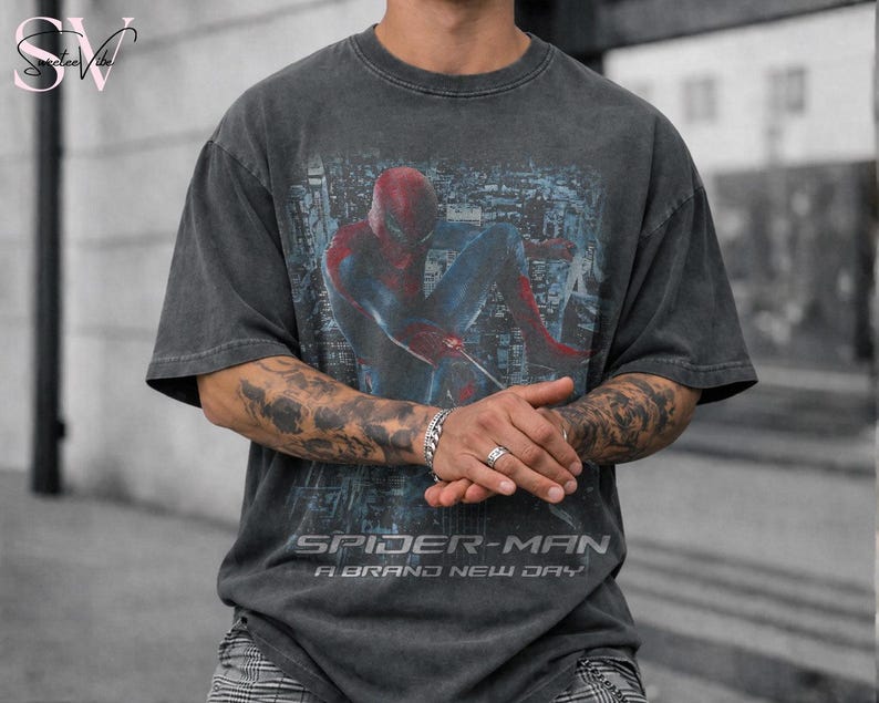 May include: A charcoal grey short-sleeve t-shirt featuring a graphic of Spider-Man in a crouched pose against a city background. The text "SPIDER-MAN A BRAND NEW DAY" is printed in white lettering below the central graphic illustration.