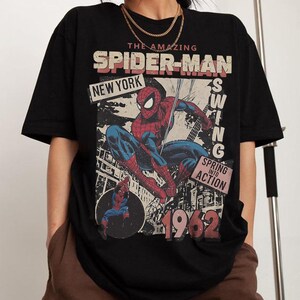 May include: Black t-shirt featuring a vintage Spider-Man graphic. The design includes the text "The Amazing Spider-Man," "New York," and "1962." The graphic shows Spider-Man in action, with red and blue colors.