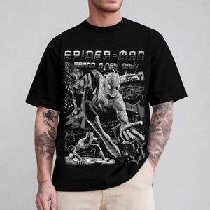May include: A black short-sleeve t-shirt featuring a graphic print of the comic book character Spider-Man in various action poses. The text "SPIDER-MAN" and "BRAND A NEW DAY" is printed in white above the black and white comic-style illustrations.