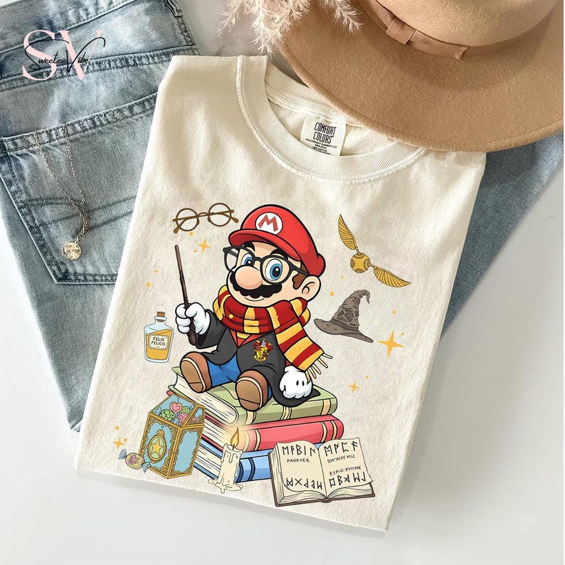 May include: Cream-colored t-shirt featuring a cartoon Mario character dressed as a wizard, complete with glasses, a wand, and a scarf. The design includes a golden snitch, a wizard hat, and potion bottles. The shirt is laid on top of blue jeans.