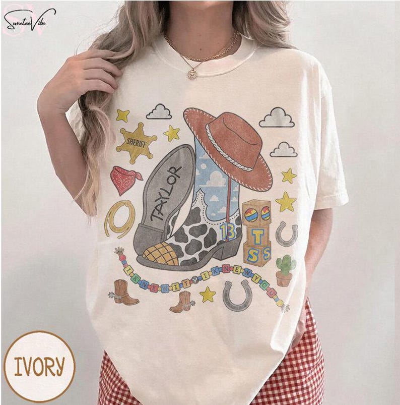 May include: An ivory t-shirt featuring a graphic print of a cowboy boot with a cow print pattern, a brown cowboy hat, a sheriff star, a horseshoe, a cactus, and wooden blocks with the letters T, S, and the number 13. The word TAYLOR is printed on the boot.