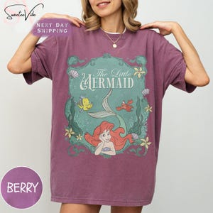 May include: Berry-colored t-shirt featuring a graphic from "The Little Mermaid." The design includes Ariel, Flounder, and Sebastian, with the words "The Little Mermaid" in a decorative font. The shirt has short sleeves and a relaxed fit.