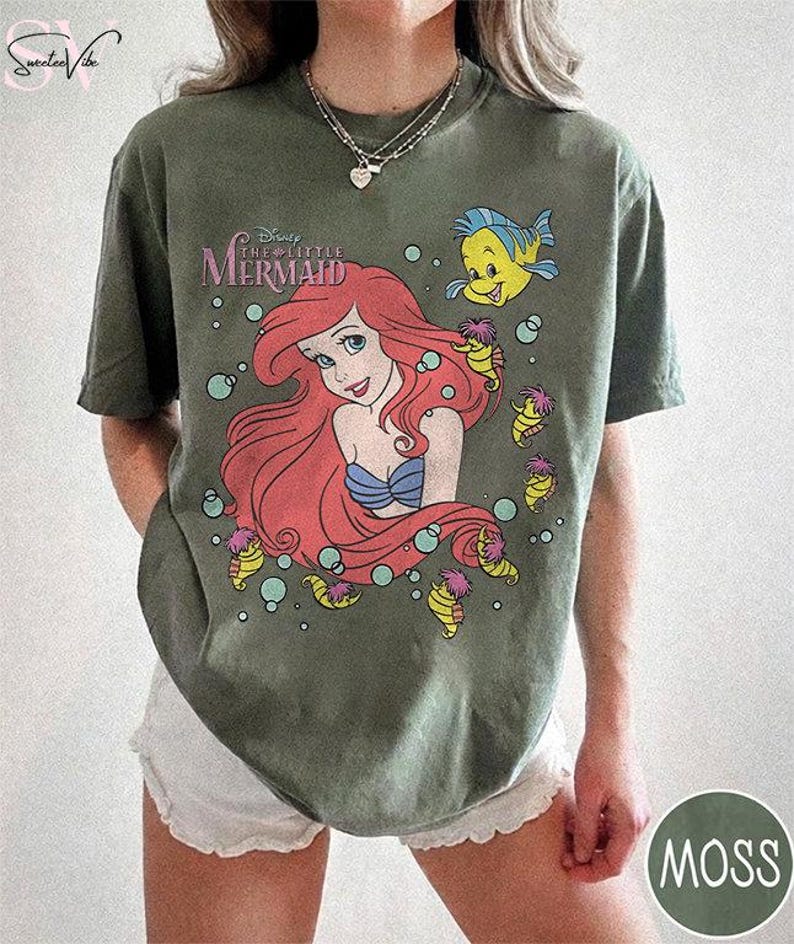 May include: Moss green t-shirt featuring a graphic of Ariel from Disney's The Little Mermaid. The design includes Ariel with red hair, a blue seashell bra, and the text "The Little Mermaid". Additional characters include a yellow fish and seahorse-like creatures.