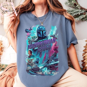 May include: A blue t-shirt featuring a graphic from the Star Wars series, "The Mandalorian and Grogu." The design includes the Mandalorian's helmet, Grogu, and other characters in a colorful, action-packed scene. Text reads "THIS IS THE WAY."