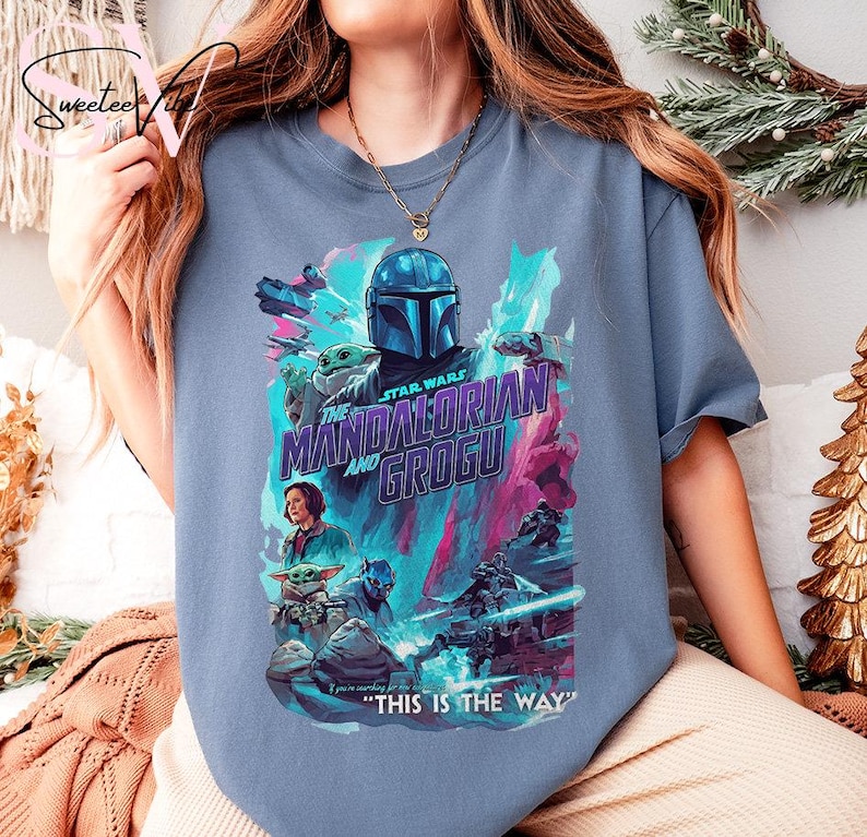May include: A blue t-shirt featuring a graphic from the Star Wars series, "The Mandalorian and Grogu." The design includes the Mandalorian's helmet, Grogu, and other characters in a colorful, action-packed scene. Text reads "THIS IS THE WAY."