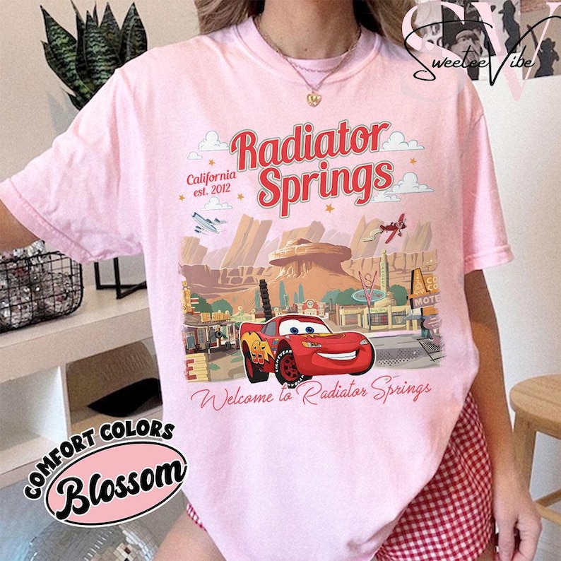 Puede incluir: Camiseta rosa claro con un gr&aacute;fico de Radiator Springs de la pel&iacute;cula Cars. El gr&aacute;fico incluye el texto "Radiator Springs" y "Welcome to Radiator Springs". La camiseta est&aacute; etiquetada como "Comfort Colors Blossom".
