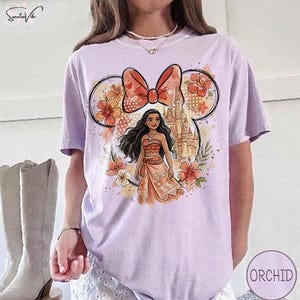 May include: A light purple t-shirt featuring a cartoon illustration of a character from a Disney movie. The design includes a bow, flowers, a castle, and the character. The shirt is a short-sleeved, casual style.