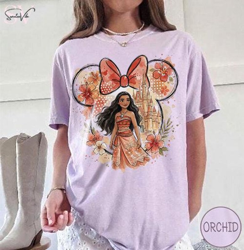 May include: A light purple t-shirt featuring a cartoon illustration of a character from a Disney movie. The design includes a bow, flowers, a castle, and the character. The shirt is a short-sleeved, casual style.