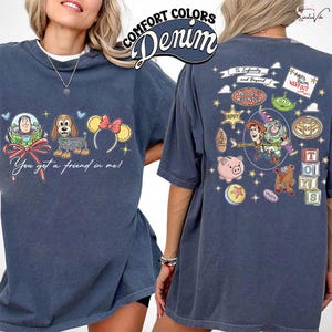 May include: A denim blue t-shirt featuring cartoon characters from Toy Story. The front displays Buzz Lightyear, a dachshund, and Minnie Mouse ears with the text "You got a friend in me!". The back has Woody, Buzz, and other characters with the text "To Infinity and Beyond".