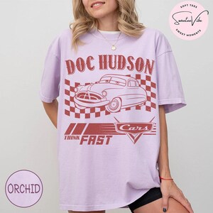 Puede incluir: Camiseta morada orqu&iacute;dea con un gr&aacute;fico rojo de Doc Hudson de la pel&iacute;cula Cars. El dise&ntilde;o incluye el texto "DOC HUDSON", un fondo a cuadros y las palabras "THINK FAST" y "Cars".