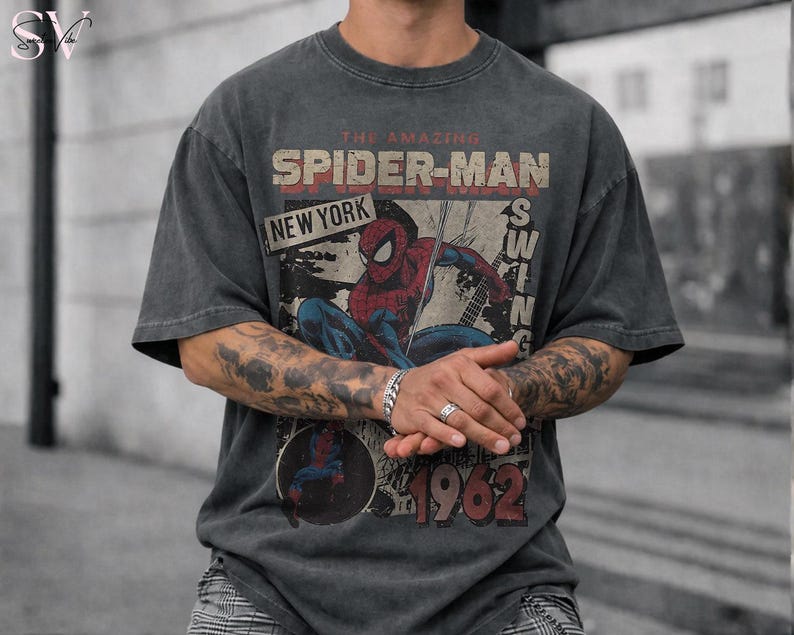 May include: Dark gray t-shirt featuring a distressed graphic of Spider-Man. The design includes the text "THE AMAZING SPIDER-MAN NEW YORK SWING 1962" in red and white. The shirt has a vintage, worn look.