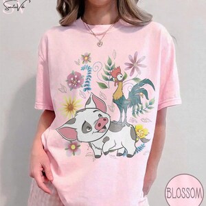 Puede incluir: Camiseta rosa claro con un dise&ntilde;o de cerdo y gallo de dibujos animados, rodeados de flores. El cerdo es blanco con manchas grises y orejas rosas. El gallo es amarillo, verde y azul. La camiseta tiene la palabra "Blossom" en un c&iacute;rculo.