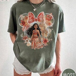 May include: Moss green t-shirt featuring a graphic of a Disney character. The design includes a castle, flowers, and a bow. The character is wearing a coral-colored outfit. The shirt is a comfortable, casual style.