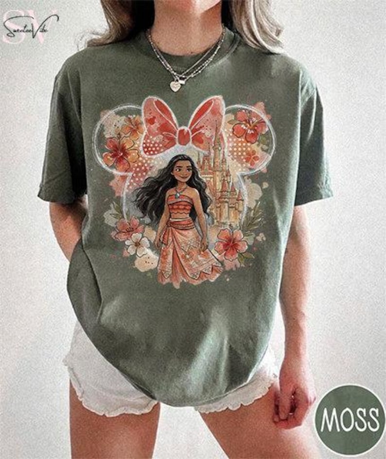 May include: Moss green t-shirt featuring a graphic of a Disney character. The design includes a castle, flowers, and a bow. The character is wearing a coral-colored outfit. The shirt is a comfortable, casual style.