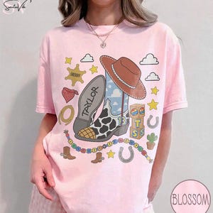 May include: A light pink t-shirt featuring a graphic print of a brown cowboy hat, a cow-print boot with the name Taylor, a sheriff badge, a red bandana, a lasso, building blocks, a cactus, horseshoes, stars, and a colorful beaded necklace.