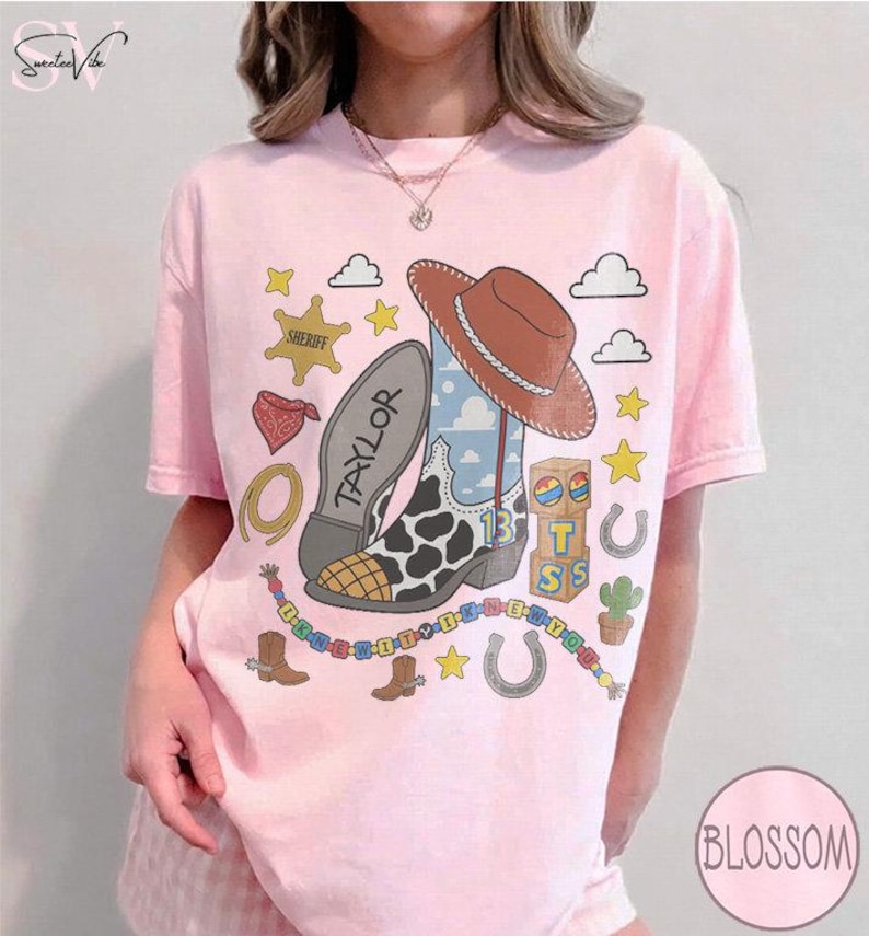 May include: A light pink t-shirt featuring a graphic print of a brown cowboy hat, a cow-print boot with the name Taylor, a sheriff badge, a red bandana, a lasso, building blocks, a cactus, horseshoes, stars, and a colorful beaded necklace.