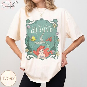 May include: Ivory-colored t-shirt featuring a teal framed graphic of Ariel from The Little Mermaid. The graphic includes the text "The Little Mermaid", a mermaid tail, and illustrations of Flounder and Sebastian. The shirt has short sleeves.