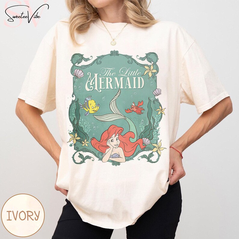 May include: Ivory-colored t-shirt featuring a teal framed graphic of Ariel from The Little Mermaid. The graphic includes the text "The Little Mermaid", a mermaid tail, and illustrations of Flounder and Sebastian. The shirt has short sleeves.