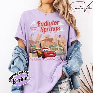 Puede incluir: Camiseta color orqu&iacute;dea con un gr&aacute;fico de Radiator Springs de la pel&iacute;cula Cars. El dise&ntilde;o incluye el texto "Radiator Springs", "California est. 2012" y "Welcome to Radiator Springs". La camiseta tambi&eacute;n tiene un gr&aacute;fico de Rayo McQueen.