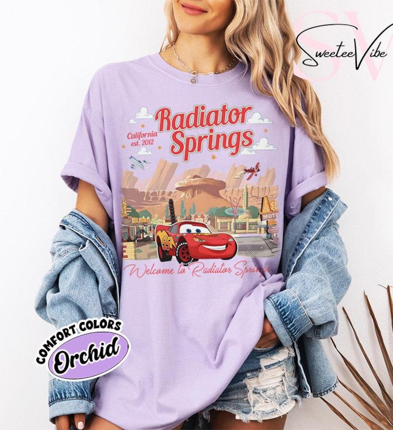 Puede incluir: Camiseta color orqu&iacute;dea con un gr&aacute;fico de Radiator Springs de la pel&iacute;cula Cars. El dise&ntilde;o incluye el texto "Radiator Springs", "California est. 2012" y "Welcome to Radiator Springs". La camiseta tambi&eacute;n tiene un gr&aacute;fico de Rayo McQueen.