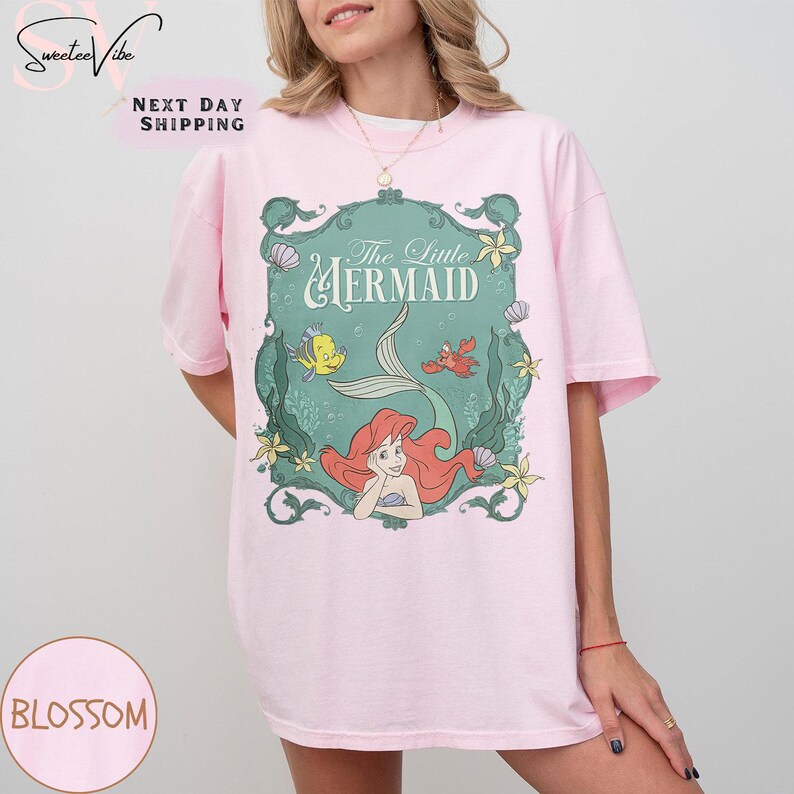 May include: A light pink t-shirt featuring a graphic of Ariel from The Little Mermaid. The graphic is framed in a teal border with the words "The Little Mermaid" and images of Flounder, Sebastian, and other sea elements.