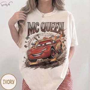 May include: Ivory-colored t-shirt featuring a graphic of Lightning McQueen from the movie Cars. The design includes the text "MC QUEEN LIGHTNING" above the car and the number "95" on the side. The shirt has a crew neck.