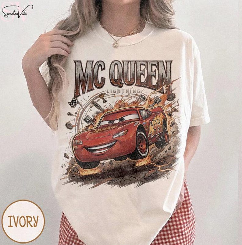 May include: Ivory-colored t-shirt featuring a graphic of Lightning McQueen from the movie Cars. The design includes the text "MC QUEEN LIGHTNING" above the car and the number "95" on the side. The shirt has a crew neck.